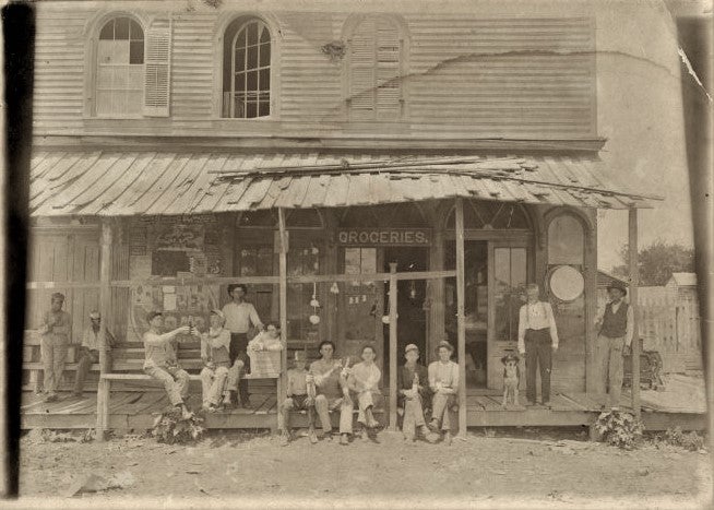 a sepia-tone photo of a run-down wooden two-story store building with several people sitting on the porch facing the photographer